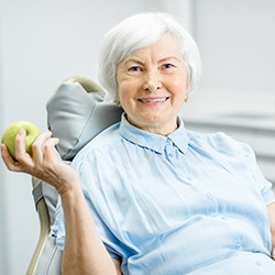 An older woman holds an apple while smiling after receiving her dental implants
