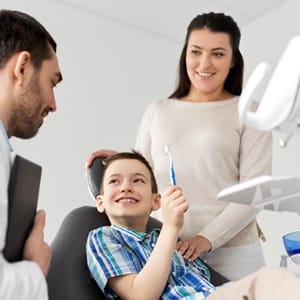 A young boy holding a toothbrush while his mother stands nearby and his dentist looks at him