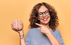 Woman with curly hair and black glasses smiling and pointing to a model brain she's holding in her other hand